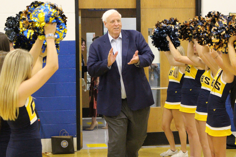 An older man in a suit walks through a hallway lined with cheerleaders in yellow uniforms, who are clapping and cheering.