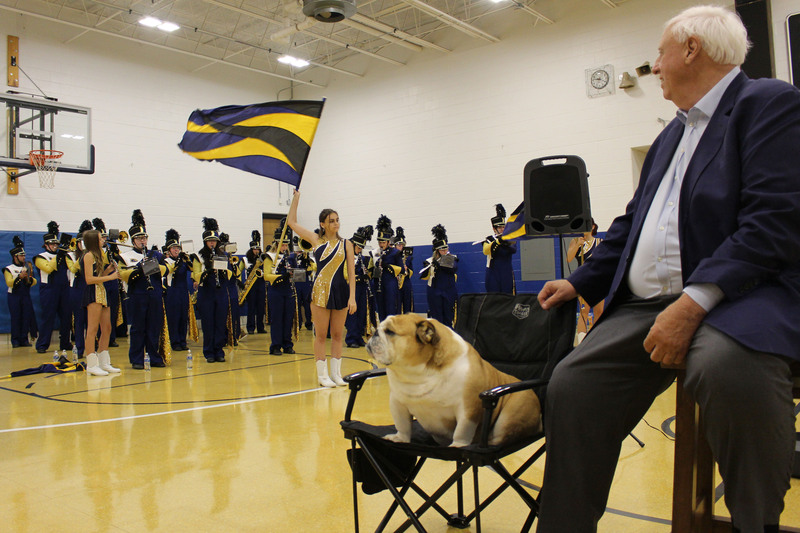 Elderly man in suit seated on chair, students in uniforms gathered behind him, basketball court and scoreboard visible.