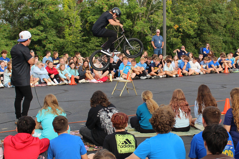 A BMX stunt performer jumps over a ramp in front of an audience of young people seated on the ground.