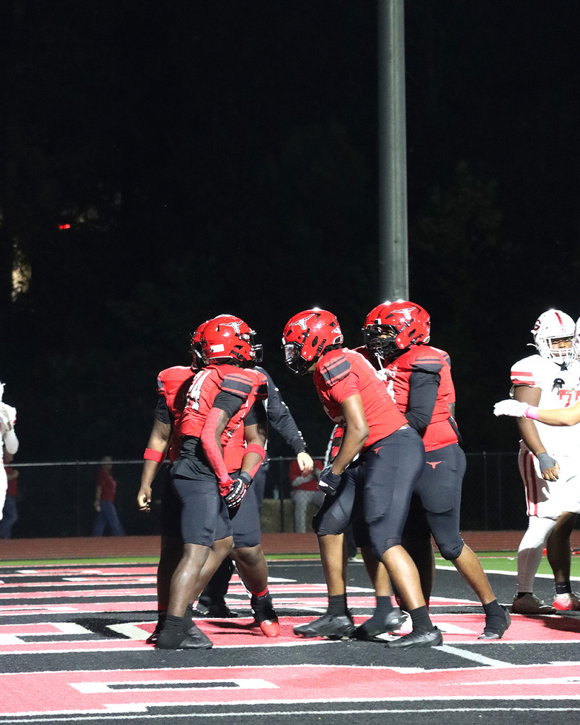 Football players in red jerseys and helmets huddle together on a field under stadium lights at night.