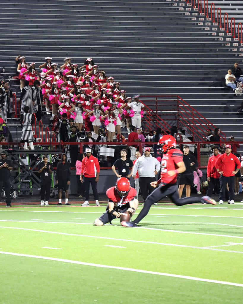 Two football players, one wearing red, one white, on the football field, one player kneeling, the other running to kick the ball. 