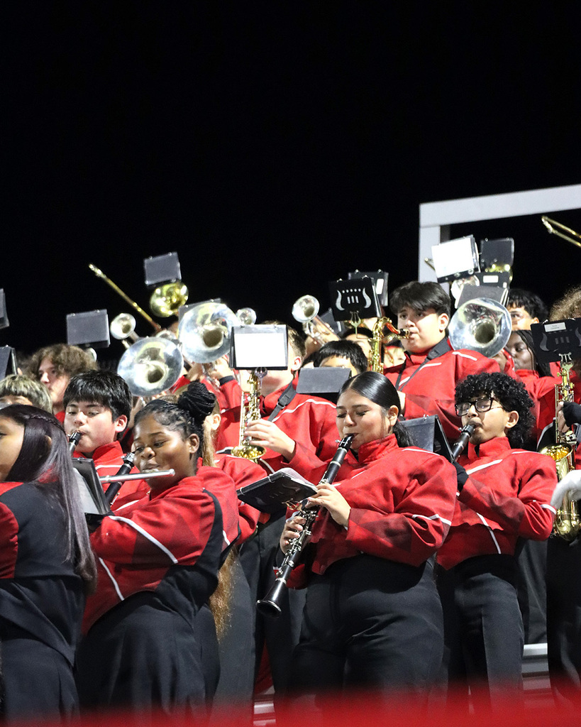 Group of band students wearing red and black uniforms playing various wind instruments in the stands.
