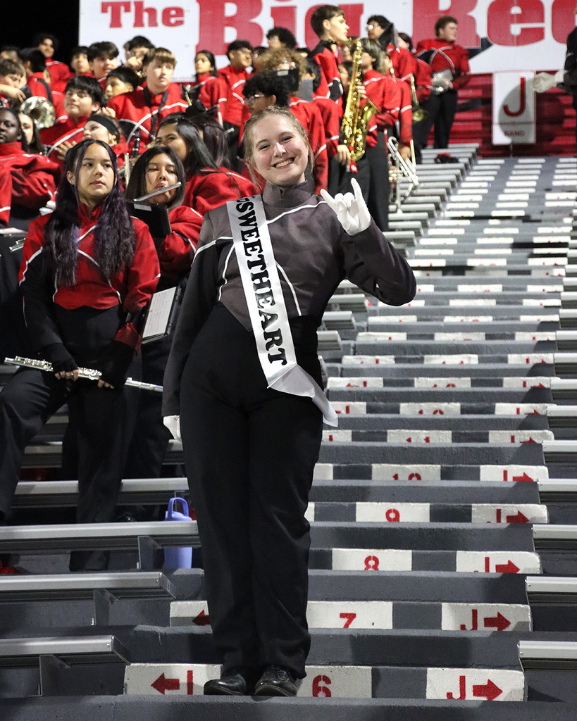 A person in a white sash stands on a bleacher, with a band in red uniforms behind.