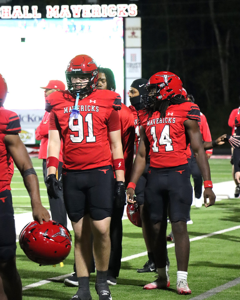 Four football players wearing red jerseys with numbers and helmets walk on a field. A scoreboard is behind them.