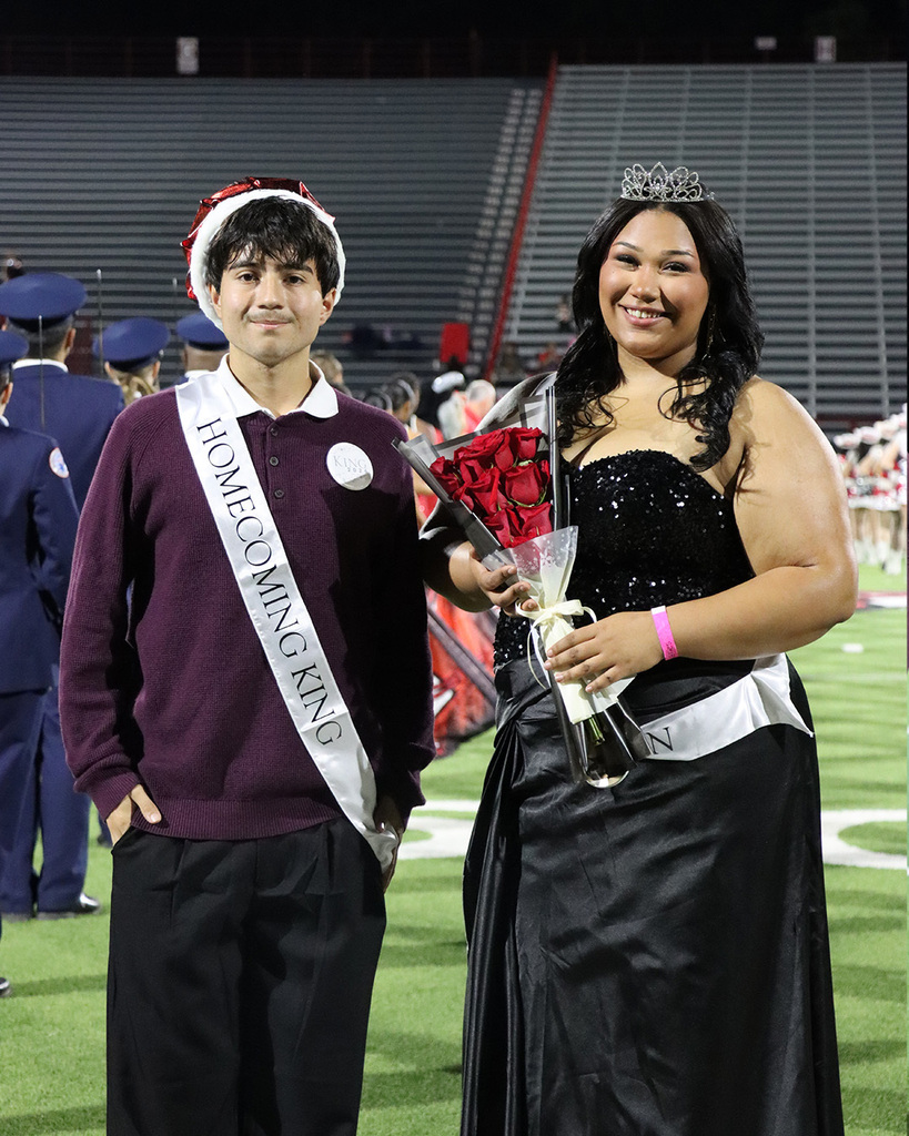A boy and girl with sashes smile, standing in an outdoor sports stadium. The boy holds roses.
