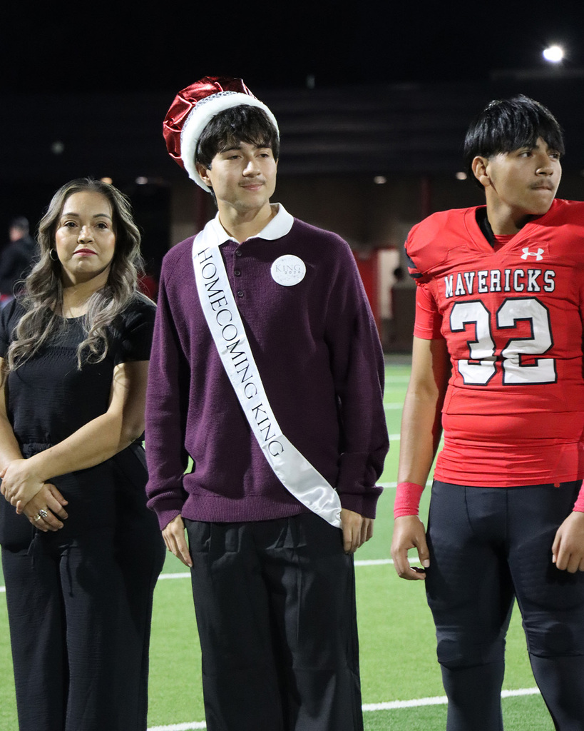 Three students standing on a football field. One wearing a homecoming sash and hat, another in a black outfit, and a third in a red football jersey.