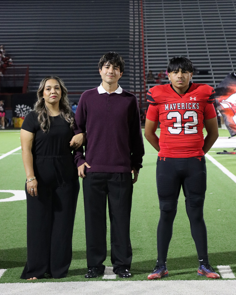 Three people stand on a field; a woman in black and two men, one in maroon and one in red football uniform.