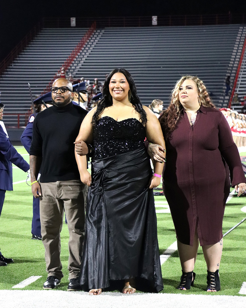 Three adults stand on a field with a bleacher background. The two women wear dresses; one has high heels. The man wears glasses and sneakers.