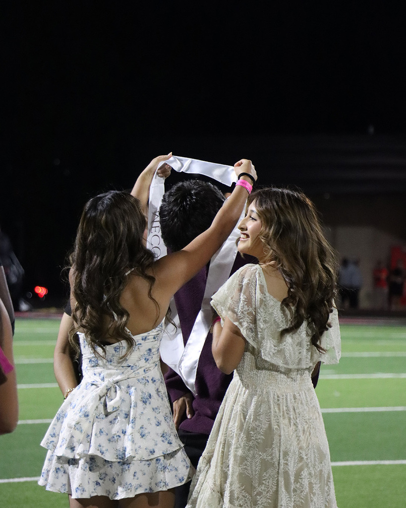 Two girls hold a sash over a person's head. One girl has a flowered dress. The other has a white dress.