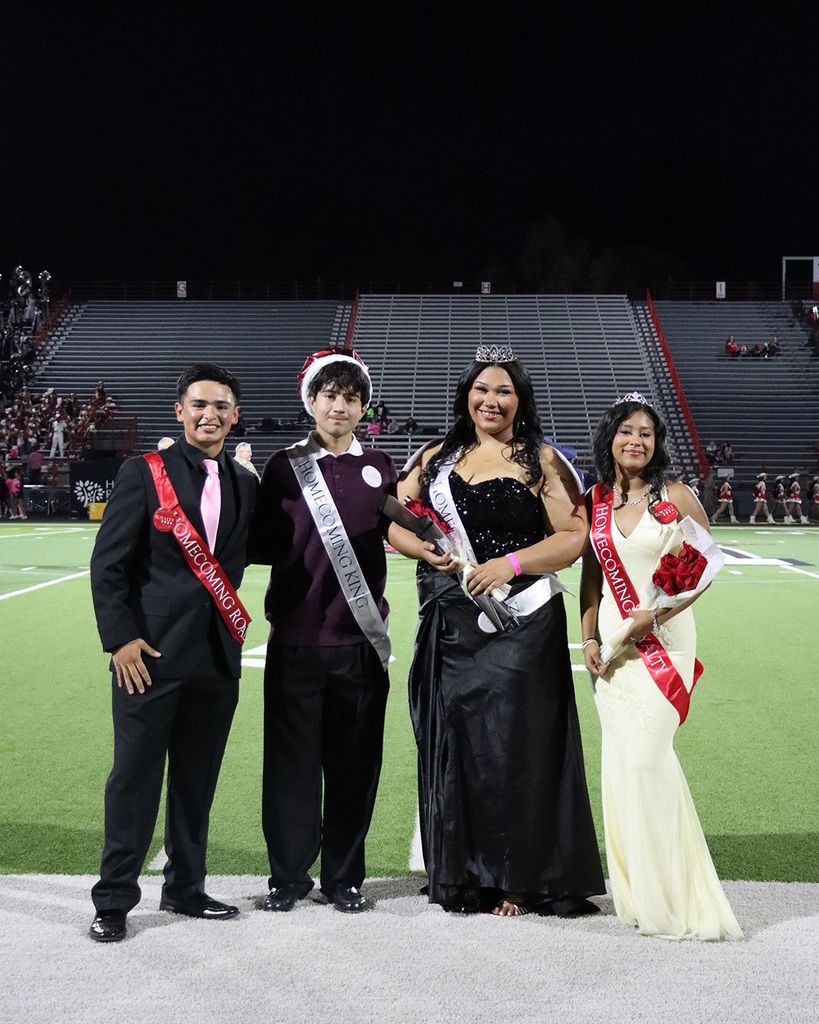 Four people stand on a football field wearing sashes. A woman holds flowers and a tiara.