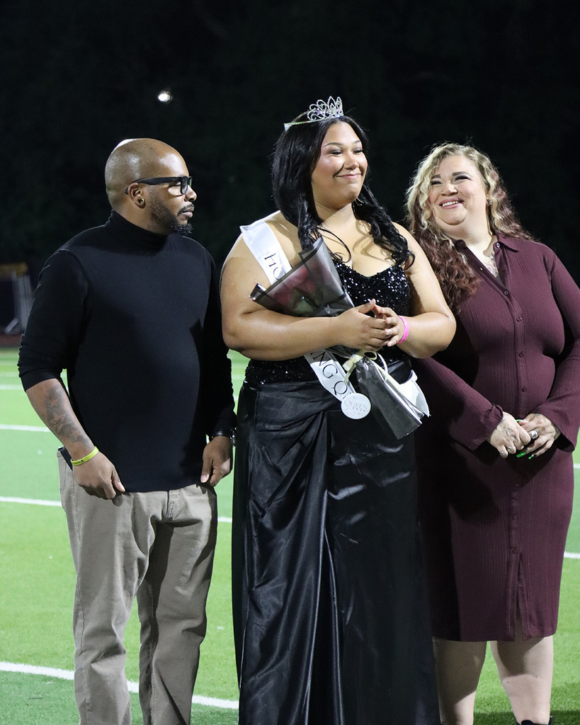 Three adults on a field, one holding a trophy, wearing a crown, and another wearing a bracelet.