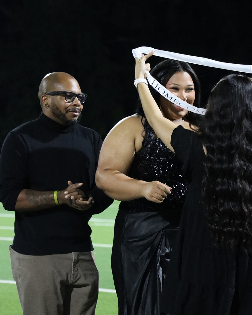 A woman holds a white sash, with a man and another woman nearby on a football field.