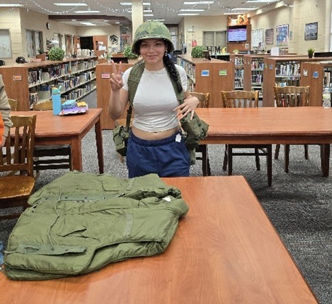 young woman wearing military backpack and hat with a military vest in front of her