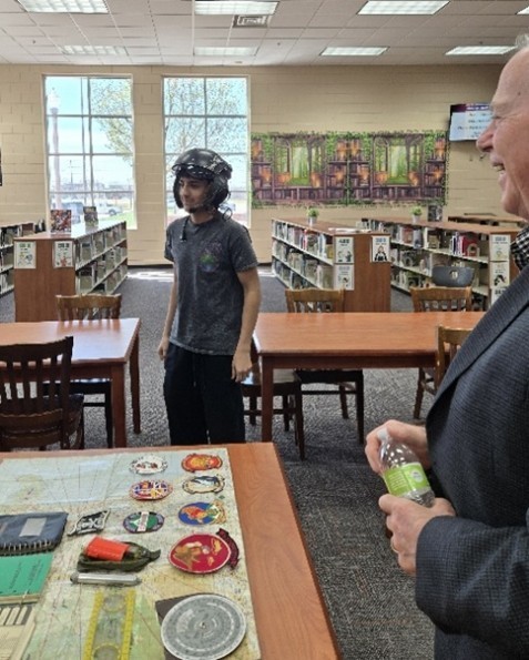 student standing with helmet with artifiacts on a table and an older gentleman in the right side of the shot.