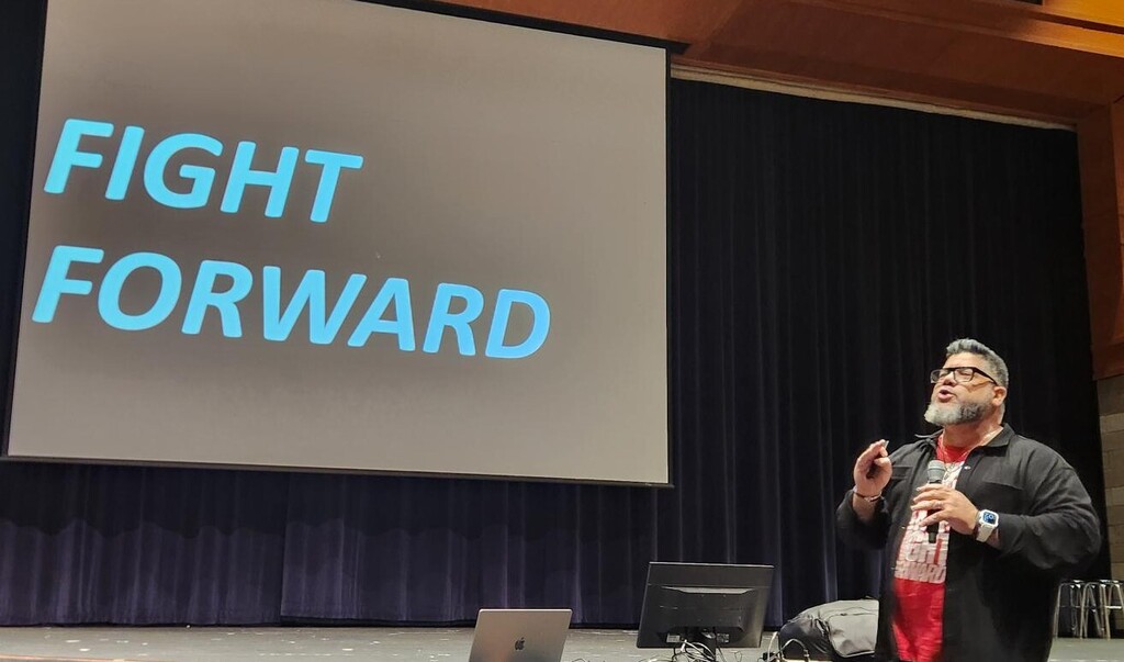 Man standing with microphone in front of powerpoint screen that says "fight forward" in blue letters.
