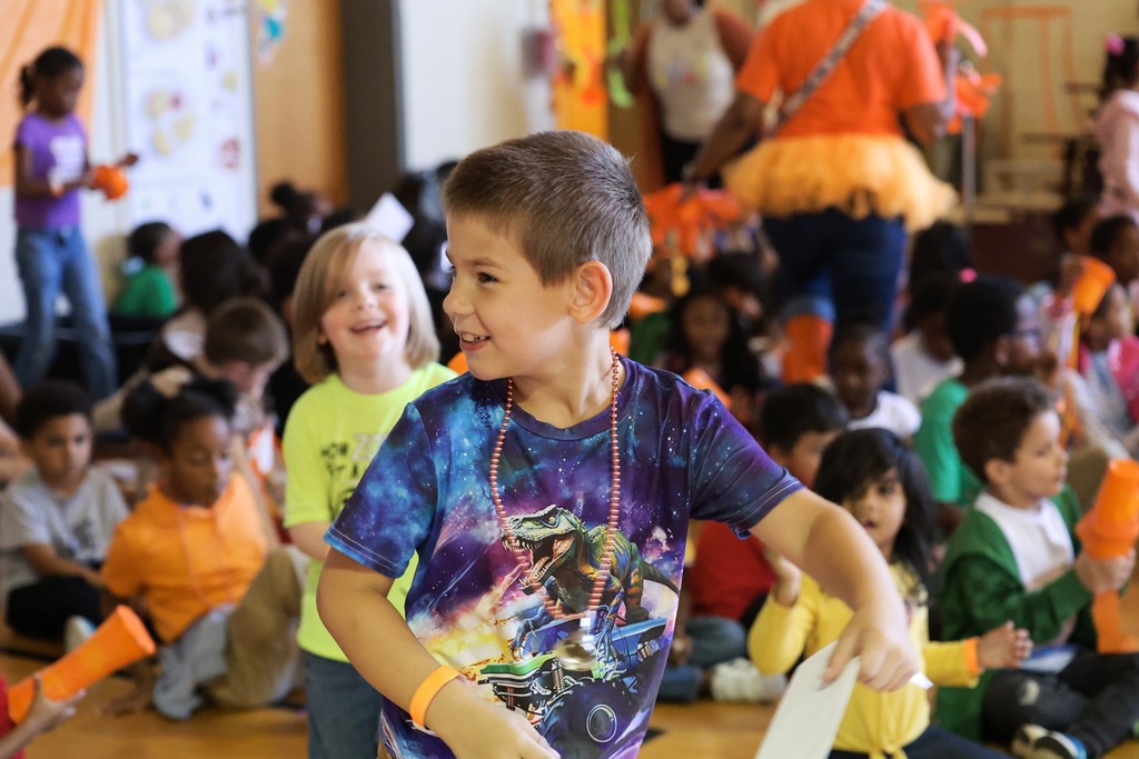 An elementary student stands smiling in the foreground. In the background are many more students seated on the gym floor.