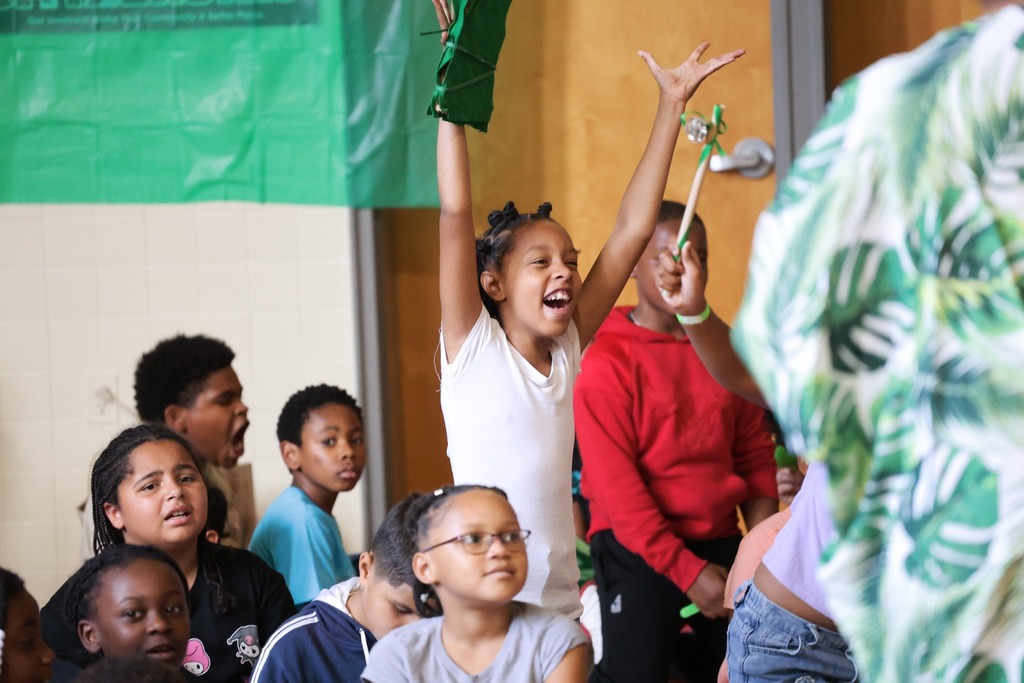 One student stands up among other seated students. Her arms are raised above her head and she smiles happily