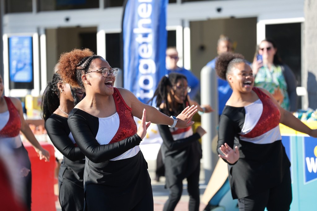 Color guard members smile and dance in front of Walmart. They wear sequined red, white, and black outfits, clapping and engaging with the crowd.