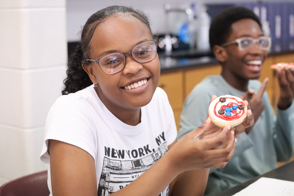 An eighth grade student smiles and holds up her completed edible atom model, made with frosting and colorful M&M’s on a cookie.