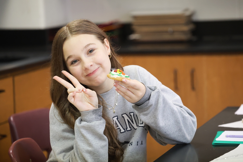 An eighth grade student smiles, holds up a peace sign, and shows her completed lithium atom model made with a frosted cookie and colorful M&M’s.