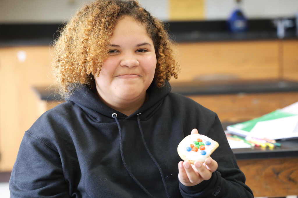 An eighth grade student smiles and holds up her completed edible atom model, made with frosting and colorful M&M’s on a cookie.