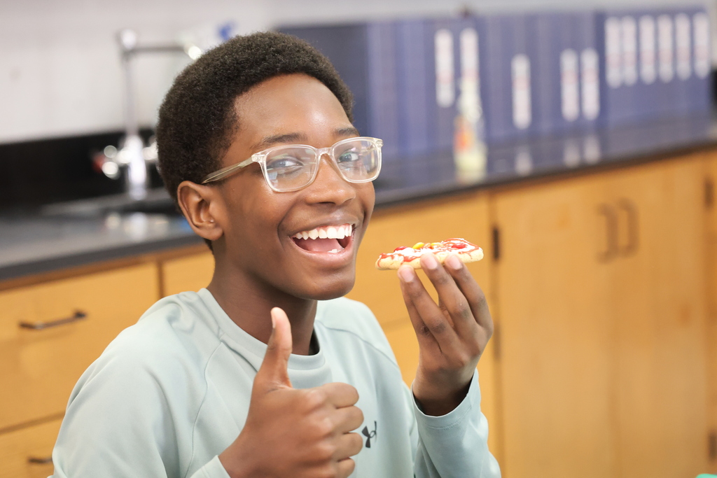 An eighth grade student smiles and gives a thumbs-up while holding his completed edible atom model made from a frosted cookie and colorful M&M’s.