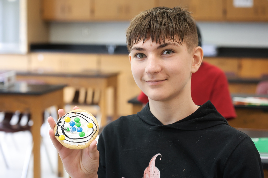 An eighth grade student smiles and holds up his completed cookie atom model, decorated with black icing and colorful M&M’s to represent parts of the atom.