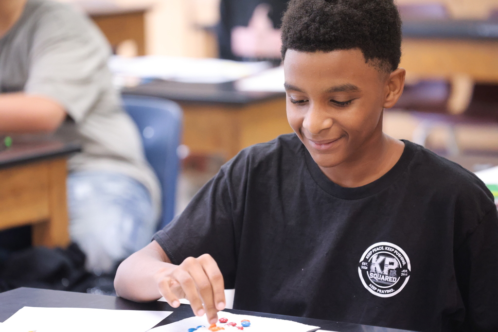 An eighth grade student smiles as he arranges colorful M&M’s during a hands-on science activity using cookies and candy to model lithium atoms.