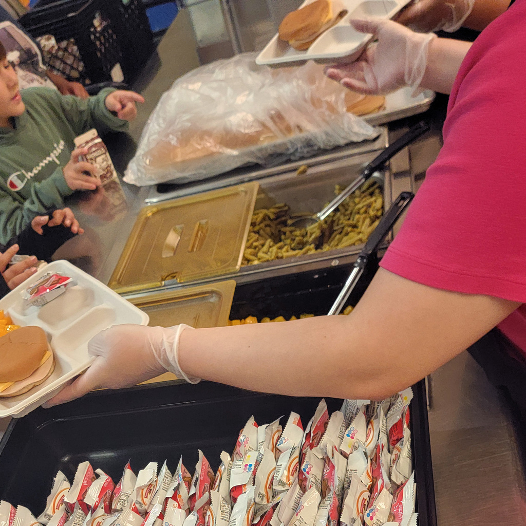 Food being served to students is seen, with one server's arm across the image. Green beans are prominent in the image