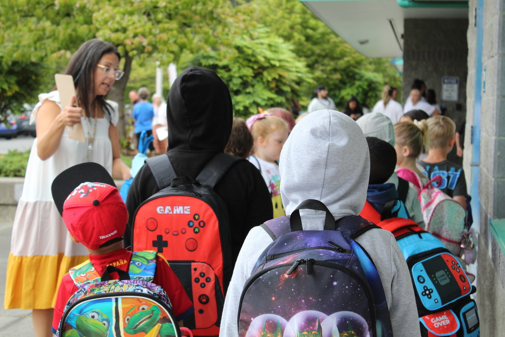 A group of children stand in line with colorful backpacks