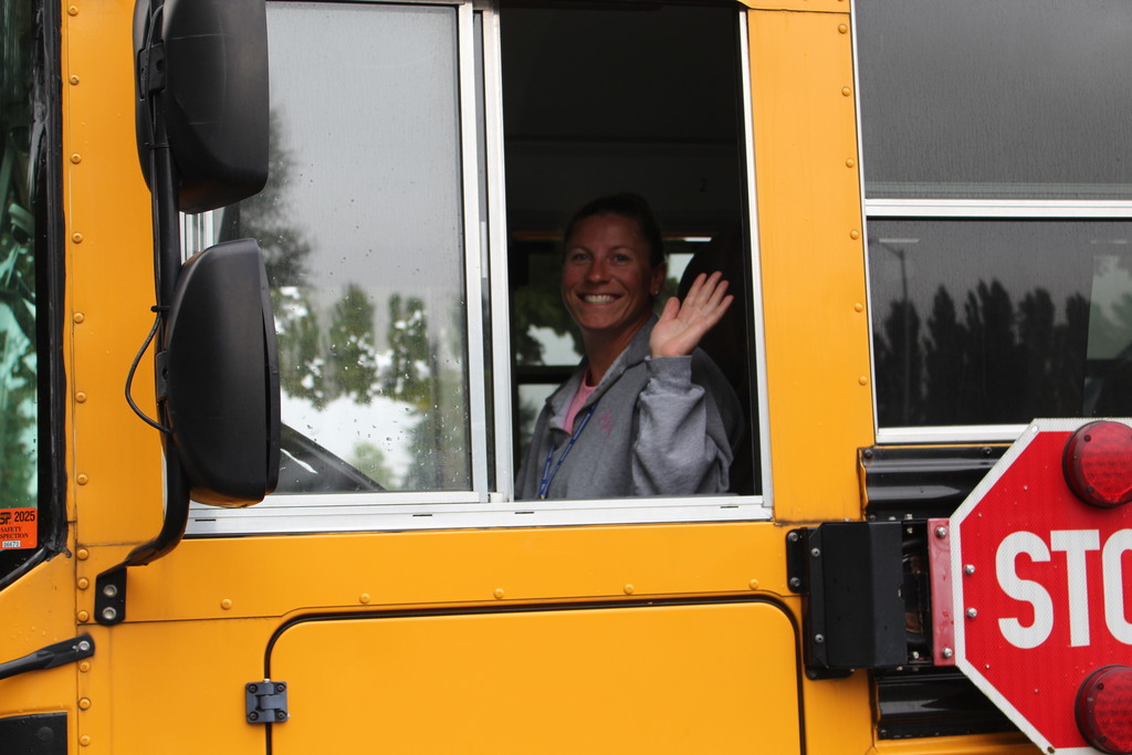 A bus driver smiles and waves from inside the stopped school bus