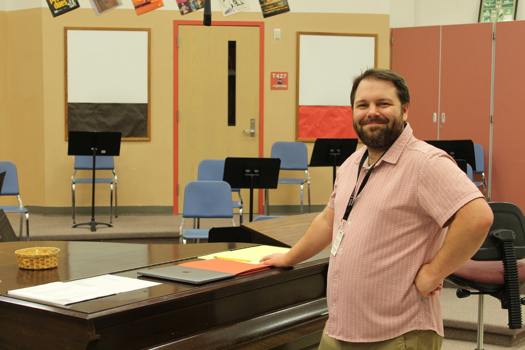 A teacher grins at the camera with music stands in the background