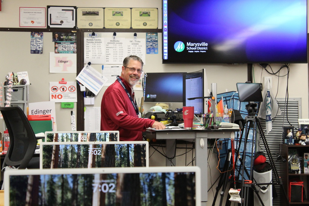 A teacher smiles at the camera in front of many computer monitors and equipment