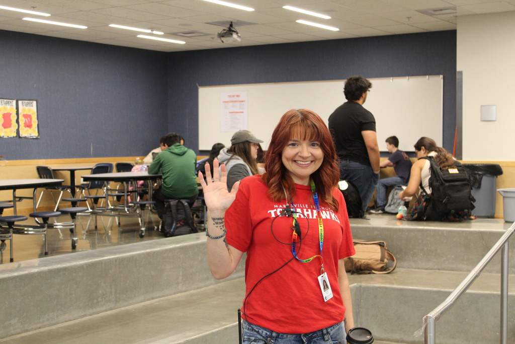 A staff member in a red shirt smiles and waves at the camera with students in the background