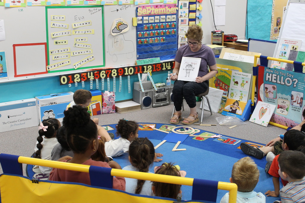 A preschool class sits in a circle and listens to their teacher while the teacher draws on a whiteboard