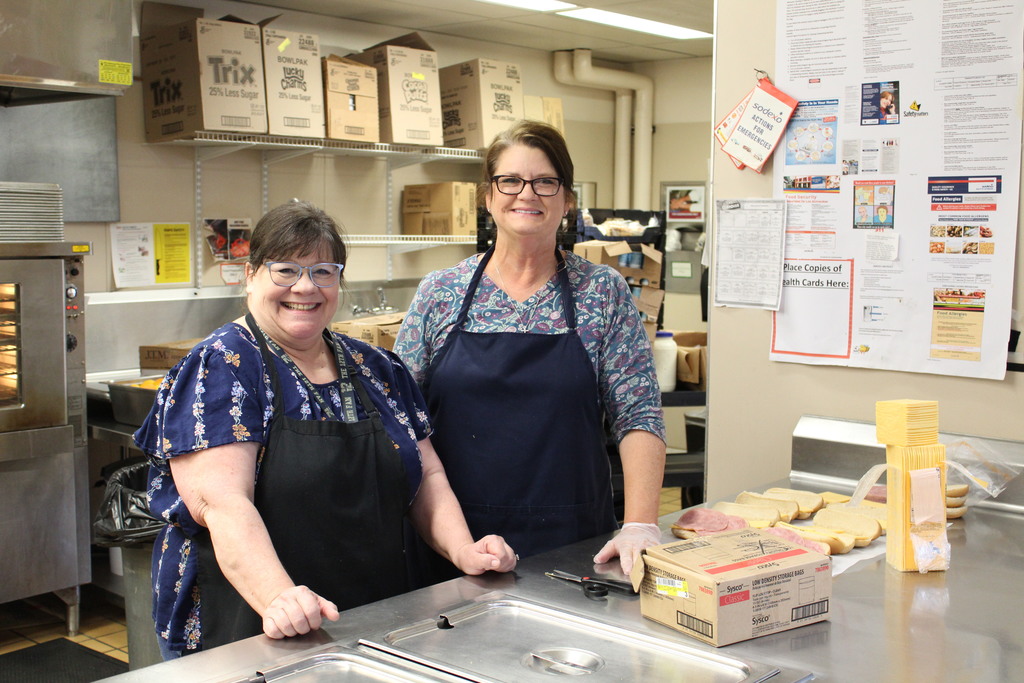 Two lunch staff smile at the camera while working in the school kitchen