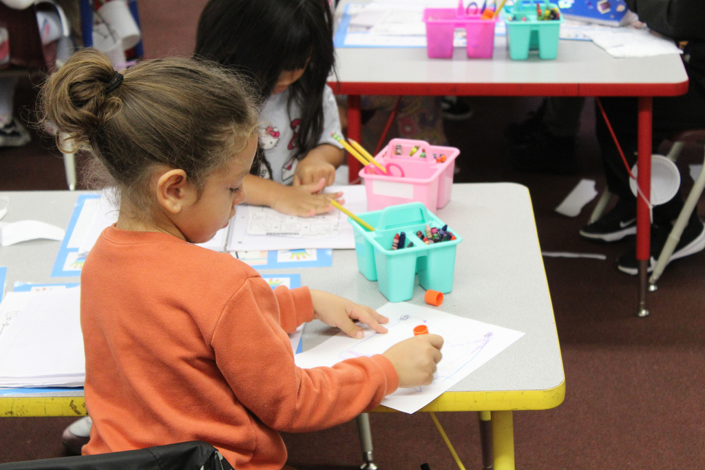 A student in an orange shirt ues a glue stick on a piece of paper while sitting at their desk