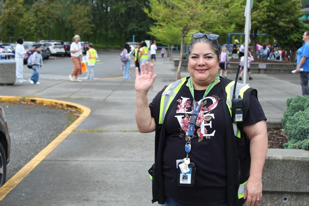 A person in a black shirt and high viz vest with a walkie talkie attached smiles and waves with trees and people in the background. 