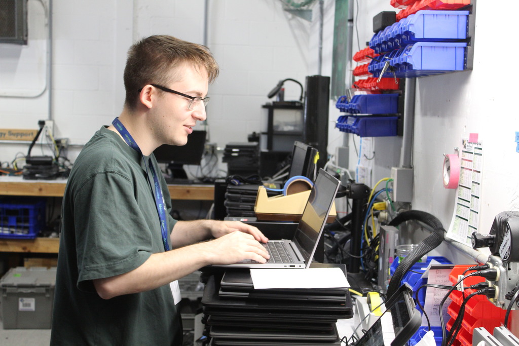 A person stands at a laptop with their hands on the keyboard, with other tech materials around him.