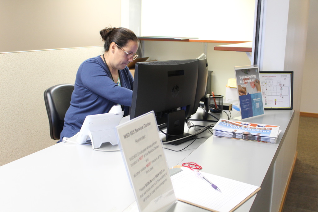 A person is seen sitting behind the counter at the MSD25 Service Center. There are some items on the counter, including a visitor sign-in sheet, newspapers, and a volunteer sign.