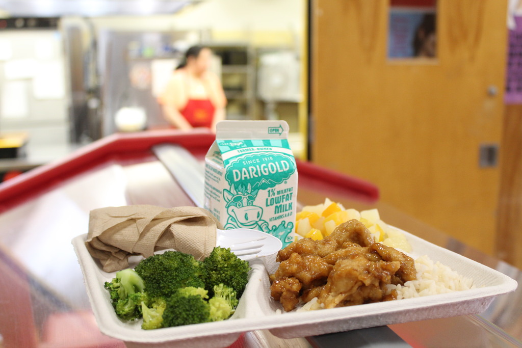 A free summer meal with fruit, broccoli, chicken, rice, and milk is seen, with a food service staff member blurry in the background.