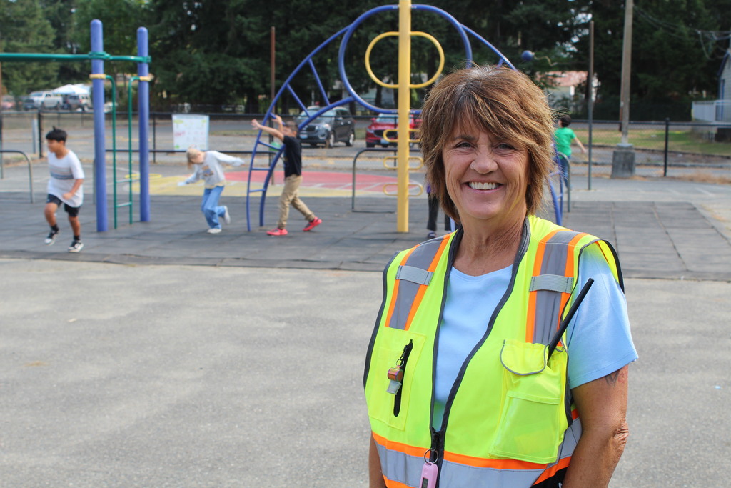 A para professional wearing a high-vis vest stands on the playground smiling at the camera while children play in the background. 