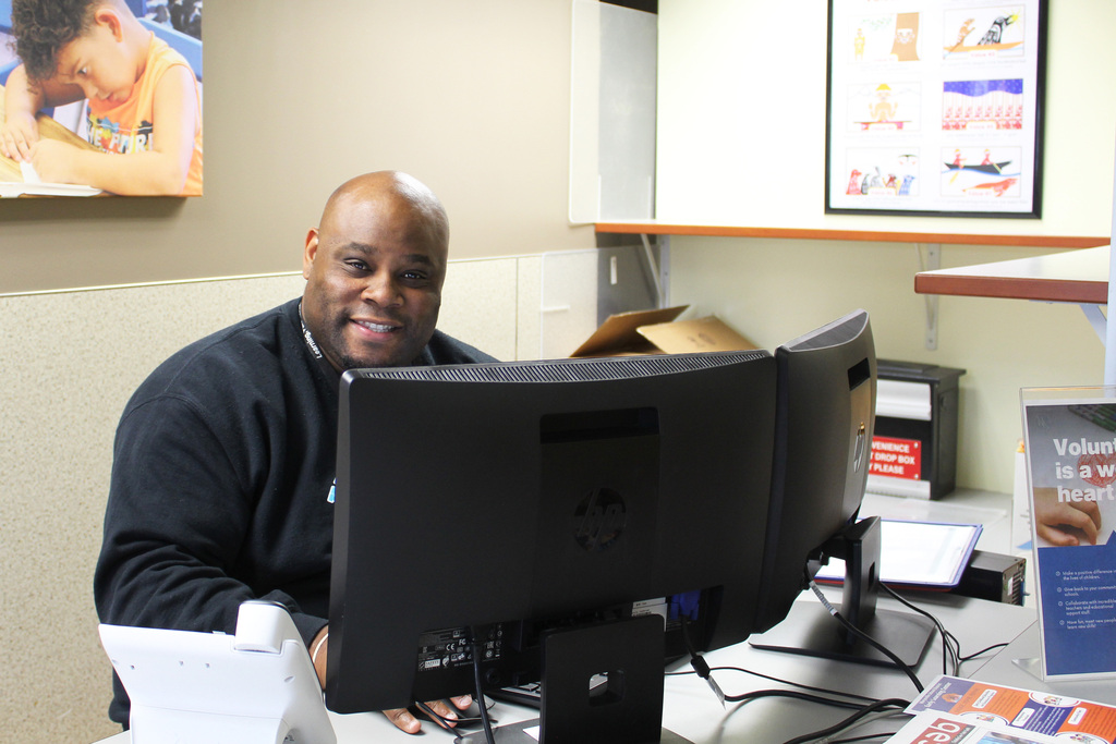 A staff member sits behind computer monitors at the Service Center front desk smiling for the camera
