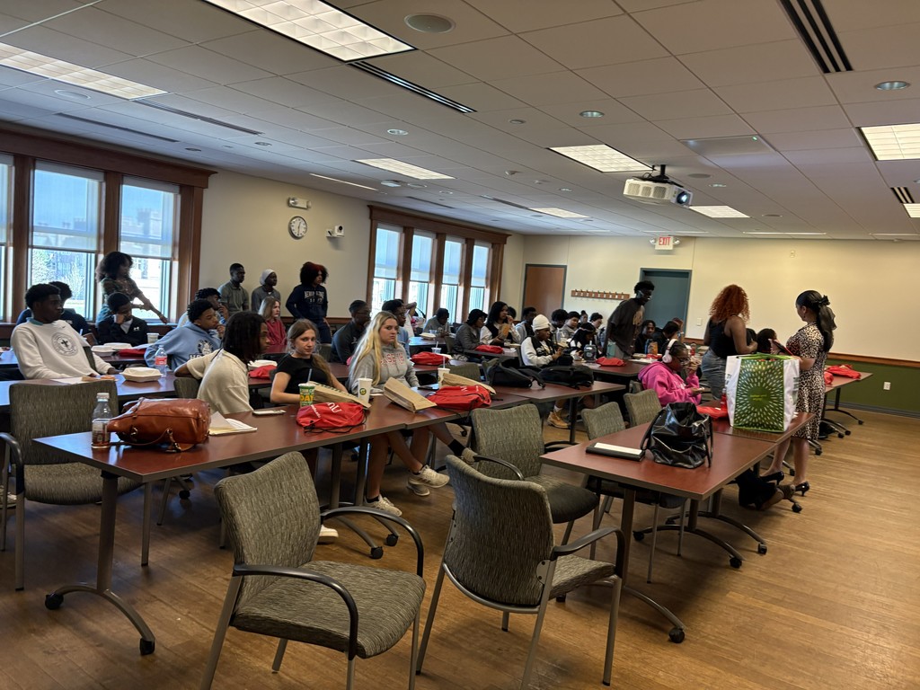 This image shows a large classroom or meeting room with many young adults gathered, likely students. They are seated and standing around long rectangular tables arranged in rows. The room has a warm, professional feel with wood-trimmed windows along the left wall letting in daylight, and a drop ceiling with fluorescent lights and a ceiling-mounted projector.  Most people appear casually dressed—hoodies, T-shirts, and jeans—and several have backpacks, notebooks, or food containers in front of them. Red fabric items, possibly T-shirts or bags, are spread across many of the tables. Some students are eating or drinking, suggesting this might be a lunch meeting or event.  At the front right side of the room, two women stand facing each other, appearing to talk or organize something. One holds or stands near a large green gift bag with a white pattern, placed on a table with a few other personal items.  The group looks relaxed and attentive, with some students facing forward and others chatting or looking around. Overall, it seems like a casual group session, workshop, or campus event in progress.  This image shows a group of about 15–18 young adults standing together on a large grassy lawn on a sunny day. They are arranged in a loose line facing the camera, smiling, as if posing for a group photo. Most appear to be college-aged and are dressed casually in T-shirts, hoodies, shorts, jeans, and sneakers.  In the center, one person holds up a colorful brochure or booklet, while another nearby holds a red drawstring bag with white text. Several others have backpacks or shoulder bags. The group appears relaxed and friendly, with some standing close together and a few slightly angled toward one another.  Behind them is a wide open campus-like setting with large, stately brick buildings featuring tall windows and decorative stonework, suggesting a university or college. The sky is clear and bright blue, and other small groups of people can be seen sitting or lying on the grass in the distance, enjoying the weather.  Overall, the scene conveys a cheerful, social moment—likely a campus gathering, tour, or group activity outdoors.  This image shows a classroom during a presentation. At the front of the room, a woman stands near a podium, facing the audience and appearing ready to speak or in the middle of presenting. She is casually dressed and positioned beside a large projection screen.  On the screen is a slide featuring a colorful book cover with a smiling young Black girl and the title “Cultivating Joyful Learning Spaces for Black Girls: Insights into Interrupting School Pushout” by Monique W. Morris. Next to the book image is a large QR code, likely for accessing more information or resources. Below, the slide includes the word “Education” and a date, March 26th, 2026.  The classroom has a modern academic feel, with a large chalkboard behind the presenter and ceiling lights overhead. In the foreground, a few students sit at round white tables. One student is working on a laptop while looking at a phone, and another sits nearby, facing slightly away. The atmosphere suggests a college lecture, workshop, or discussion focused on education and equity.