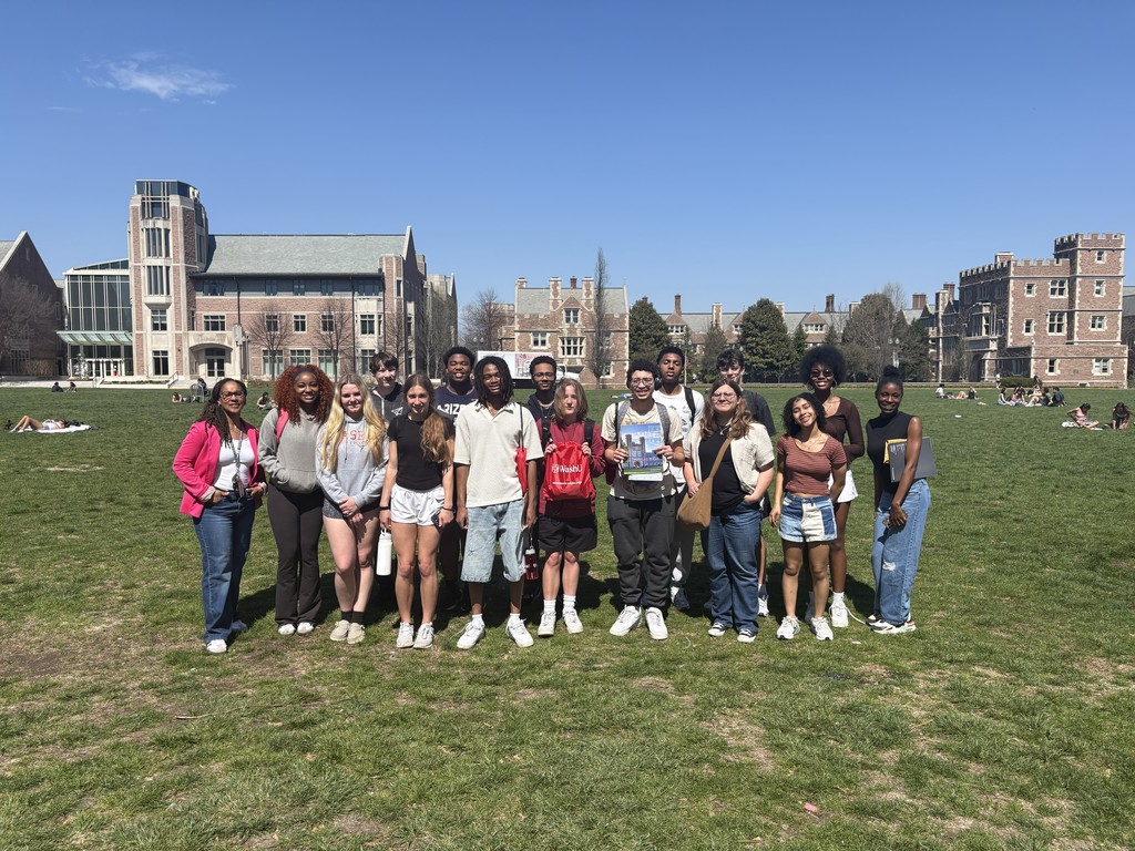 This image shows a group of about 15–18 young adults standing together on a large grassy lawn on a sunny day. They are arranged in a loose line facing the camera, smiling, as if posing for a group photo. Most appear to be college-aged and are dressed casually in T-shirts, hoodies, shorts, jeans, and sneakers.  In the center, one person holds up a colorful brochure or booklet, while another nearby holds a red drawstring bag with white text. Several others have backpacks or shoulder bags. The group appears relaxed and friendly, with some standing close together and a few slightly angled toward one another.  Behind them is a wide open campus-like setting with large, stately brick buildings featuring tall windows and decorative stonework, suggesting a university or college. The sky is clear and bright blue, and other small groups of people can be seen sitting or lying on the grass in the distance, enjoying the weather.  Overall, the scene conveys a cheerful, social moment—likely a campus gathering, tour, or group activity outdoors.