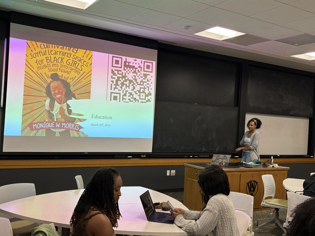 This image shows a classroom during a presentation. At the front of the room, a woman stands near a podium, facing the audience and appearing ready to speak or in the middle of presenting. She is casually dressed and positioned beside a large projection screen.  On the screen is a slide featuring a colorful book cover with a smiling young Black girl and the title “Cultivating Joyful Learning Spaces for Black Girls: Insights into Interrupting School Pushout” by Monique W. Morris. Next to the book image is a large QR code, likely for accessing more information or resources. Below, the slide includes the word “Education” and a date, March 26th, 2026.  The classroom has a modern academic feel, with a large chalkboard behind the presenter and ceiling lights overhead. In the foreground, a few students sit at round white tables. One student is working on a laptop while looking at a phone, and another sits nearby, facing slightly away. The atmosphere suggests a college lecture, workshop, or discussion focused on education and equity.