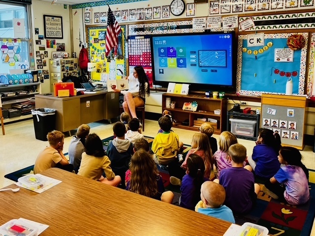 Picture of a student reading to a group of elementary students that are sitting on the floor gathered around her. 
