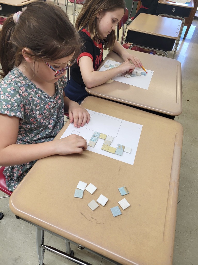 Students in a 3rd grade classroom working on math activities involving area and perimeter, using shapes and hands-on materials.