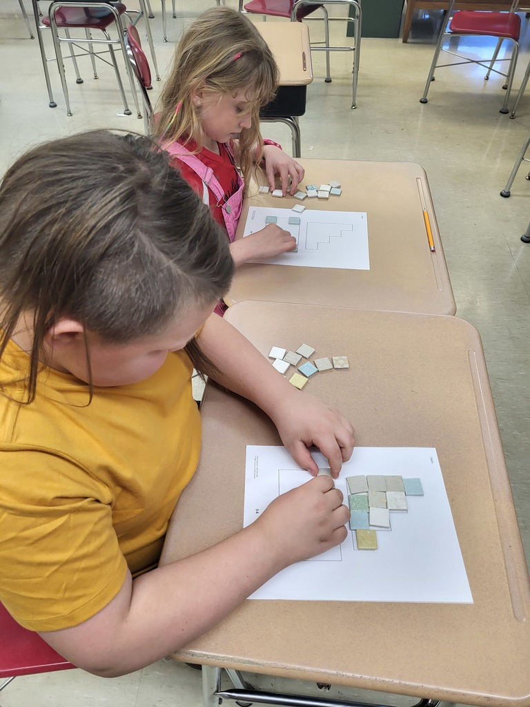 Students in a 3rd grade classroom working on math activities involving area and perimeter, using shapes and hands-on materials.