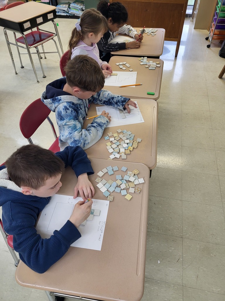Students in a 3rd grade classroom working on math activities involving area and perimeter, using shapes and hands-on materials.